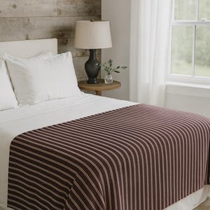 May include: A bed with white bedding and a burgundy and white striped bed runner. A dark lamp sits on a small wooden side table. The background features a wooden wall and a window with white curtains.