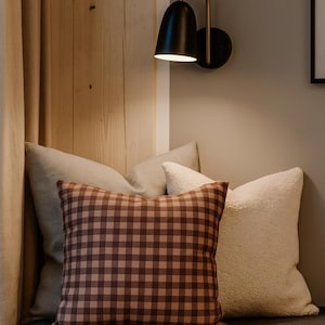 May include: A cozy seating area with a brown and tan gingham patterned pillow, a cream-colored fluffy pillow, and a light-colored pillow. A black and gold wall sconce provides soft lighting. The pillows rest on a gray cushion.