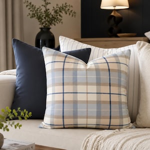 May include: A collection of decorative throw pillows in various patterns and colors on a beige sofa. The pillows include a navy blue solid pillow, a plaid pillow with blue and beige tones, and a striped pillow.