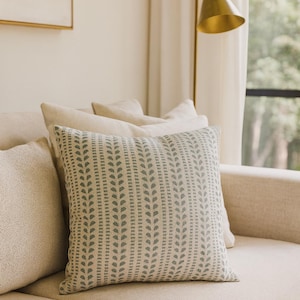 May include: A beige sofa with several decorative cushions. The central cushion has a repeating pattern of vertical rows of small, light green leaf shapes on a beige background. A gold-coloured lamp is in the background.