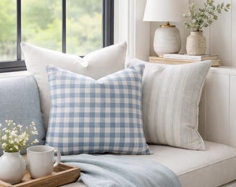 Op de afbeelding: Een verzameling decoratieve sierkussens, waaronder een kussen met een blauw-wit vichy-ruitpatroon, een effen gebroken wit kussen en een kussen met blauwe verticale strepen, vergezeld door een lichtblauwe plaid met franjes op een bank.