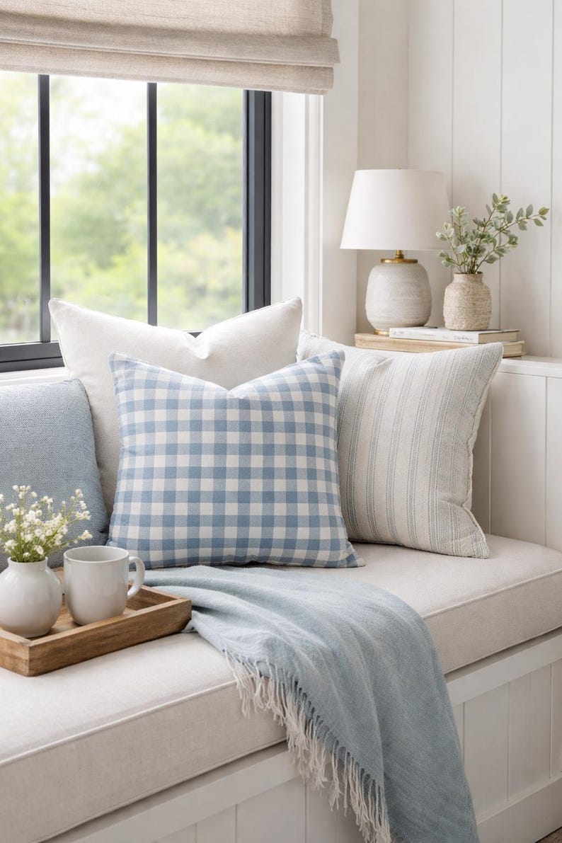 Pu&ograve; includere: Una collezione di cuscini decorativi che include un modello con motivo a quadretti vichy blu e bianchi, un cuscino color bianco sporco a tinta unita e uno con strisce verticali blu, accompagnati da una coperta azzurra con frange su una panca.