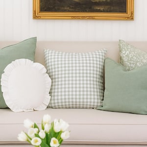 Puede incluir: Un sofá beige con varios cojines decorativos en tonos verdes y blancos. Una almohada redonda blanca con borde con volantes está al lado de una almohada de cuadros. Un jarrón de tulipanes blancos está sobre una mesa tejida.