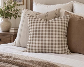 May include: A square throw pillow featuring a brown and white gingham check pattern. The pillow is positioned in the center of a bed, layered in front of a larger beige pillow with vertical stripes and a solid brown pillow on a white bedspread.