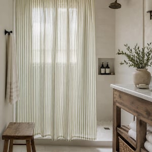 May include: A shower curtain featuring a vertical striped pattern in sage green and white. The curtain hangs from a black rod with rings, covering a shower area. The fabric has a light, semi-sheer appearance with consistent vertical lines throughout.