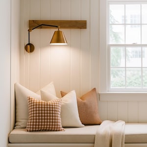 May include: A comfortable reading nook featuring a brass wall lamp, wooden detail, and a bench with cushions and a blanket. The cushions include a brown gingham pattern and neutral colours. The window provides natural light.