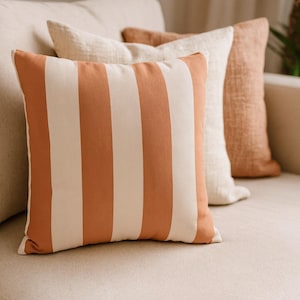 Puede incluir: Tres cojines decorativos sobre un sofá beige. El cojín delantero tiene rayas verticales en terracota y crema. Los otros dos cojines son lisos en crema y terracota. Los cojines añaden un toque de color y textura al sofá de tonos neutros.
