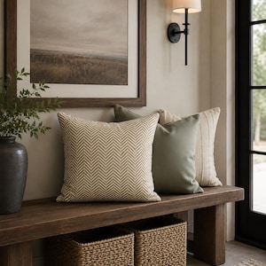 May include: A wooden bench with three decorative pillows in varying shades of green and beige. Two woven baskets sit below the bench. A framed landscape painting and a wall sconce are visible in the background.