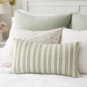 Könnte beinhalten: Ein Bett mit mehreren dekorativen Kissen. Die Kissen umfassen ein gestreiftes Lendenkissen mit grünen und weißen Streifen, Kissen mit Blumendruck und einfarbige grüne und weiße Kissen. Das Bett ist mit einer weißen Bettdecke bedeckt.
