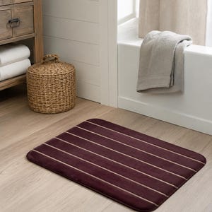 May include: A rectangular bath mat featuring a deep burgundy color with thin, horizontal beige stripes. The mat has rounded corners and a soft, plush texture, providing a simple and functional design for a bathroom floor space.