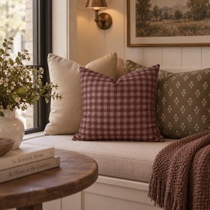 May include: A square throw pillow featuring a dark red and white gingham check pattern, positioned on a window seat alongside a solid beige pillow and an olive green pillow with a small white floral print, accompanied by a textured dark red throw.