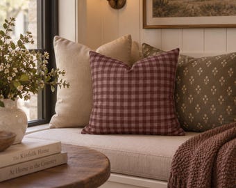 May include: A square throw pillow featuring a dark red and white gingham check pattern, positioned on a window seat alongside a solid beige pillow and an olive green pillow with a small white floral print, accompanied by a textured dark red throw.