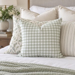 Op de afbeelding: Een vierkant sierkussen met een saliegroen en wit vichy-ruitpatroon. Het kussen is geplaatst tussen andere sierkussens op een bed, waaronder een met een botanische print en een ander met verticale strepen in neutrale tinten.