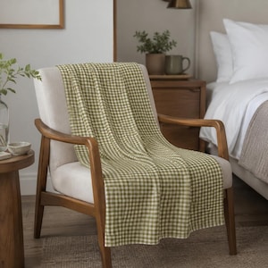 May include: A rectangular throw blanket featuring a green and white gingham check pattern. The fabric has a visible woven texture and is draped over the seat and backrest of a light-colored upholstered armchair with wooden armrests and legs.