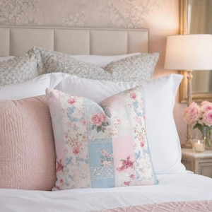 May include: A bed with several pillows, including a patchwork pillow with floral patterns in pink and blue. Other pillows are solid pink and patterned with a blue floral design. The bed has a white comforter and a pink textured blanket.