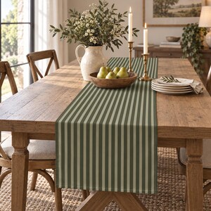 May include: A rectangular table runner featuring a vertical striped pattern in alternating shades of sage green and dark olive green. The fabric runner is laid flat across the center of a wooden dining table, extending from one end to the other.