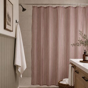 May include: A shower curtain featuring a vertical striped pattern in muted red and white. The curtain hangs from a silver metal rod with circular rings, extending across the width of the shower area to provide coverage for the tiled interior space.