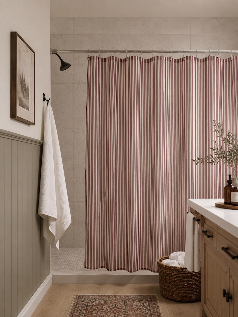 May include: A shower curtain featuring a vertical striped pattern in muted red and white. The curtain hangs from a silver metal rod with circular rings, extending across the width of the shower area to provide coverage for the tiled interior space.