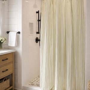 May include: A shower curtain featuring a vertical striped pattern in light sage green and white. The curtain hangs from a silver metal rod with circular rings, covering the opening of a walk-in shower stall with white subway tile walls.