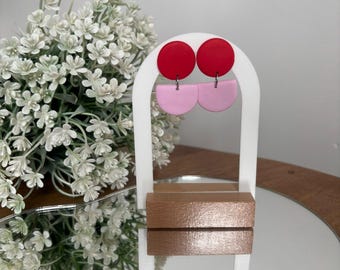 May include: A white earring display stand with a wooden base holds two pairs of earrings. Each pair features a red circle and a pink semi-circle. The earrings are displayed against a mirrored surface, with white flowers in the background.