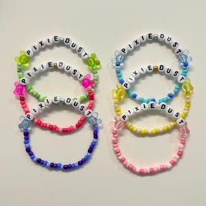 May include: Six colorful beaded bracelets with the words "PIXIE DUST" spelled out in black letters on white beads. The bracelets are in shades of pink, red, blue, green, and yellow, with decorative charms.