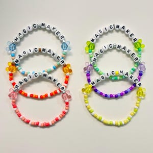 May include: Colorful beaded bracelets with the words "MAGIC MAKER" spelled out in white letter beads. The bracelets feature various colors, including red, orange, pink, blue, green, and yellow, with flower-shaped accents.