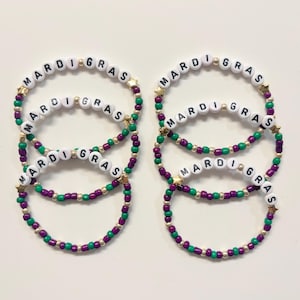 May include: Several beaded bracelets in Mardi Gras colors. The bracelets are purple, green, and gold, with the words "MARDI GRAS" spelled out in white letter beads. Gold star-shaped beads are interspersed throughout.