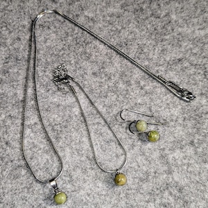 Könnte beinhalten: Ein Set aus silbernen Kettenhalsbändern mit grünen Steinanhängern und passenden Ohrringen. Die Halsbänder sind in verschiedenen Längen und die Steine sind rund.