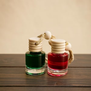 May include: Two small glass bottles with wooden caps and rope hangers. One bottle contains green liquid, the other red. The bottles sit on a dark wooden surface against a neutral background. The caps are light-colored and cylindrical.