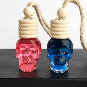 May include: Two skull-shaped car air fresheners, one red and one blue, with wooden tops and a rope for hanging. The glass bottles have a detailed skull design. The background is a dark surface.