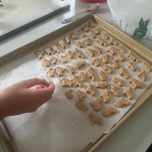 May include: A baking sheet lined with parchment paper and filled with various animal-shaped cookies. The cookies are a light brown color and have a slightly rough texture. A hand is reaching towards the cookies, as if to pick one up.