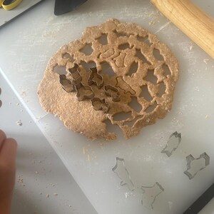 May include: A white cutting board with a rolling pin, a spatula, and a sheet of dough with various cookie cutters. The dough has been cut into shapes using the cookie cutters. There are also several cookie cutters on the cutting board.