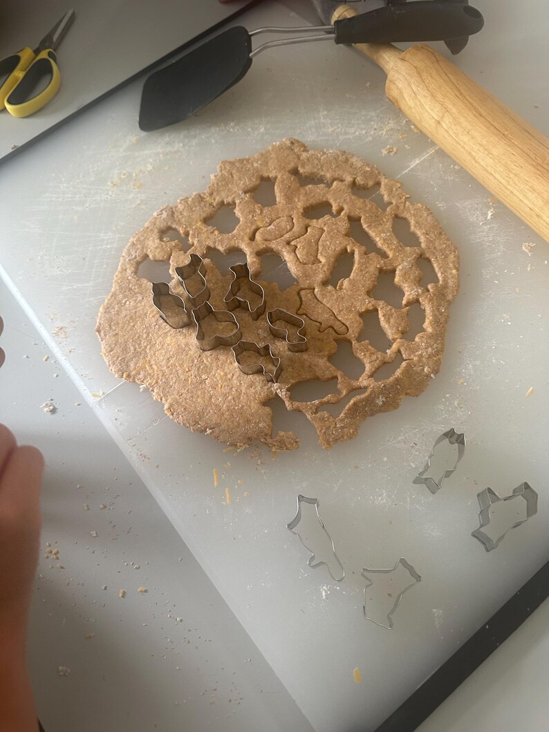 May include: A white cutting board with a rolling pin, a spatula, and a sheet of dough with various cookie cutters. The dough has been cut into shapes using the cookie cutters. There are also several cookie cutters on the cutting board.