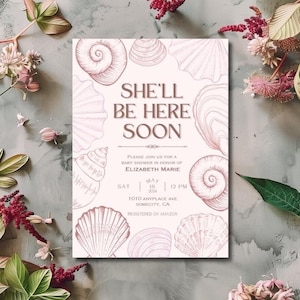 Puede incluir: Una invitación de baby shower rosa y blanca con un diseño de conchas marinas. El texto dice "She'll Be Here Soon" e incluye la fecha, la hora y la ubicación del evento. La invitación también indica que el evento es en honor de Elizabeth Marie y que los regalos están registrados en Amazon.
