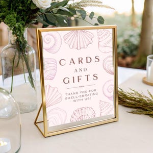 Może przedstawiać: Złota ramka z napisem "CARDS AND GIFTS" w brązowych literach. Znak ma jasnoróżowe tło z ilustracjami muszli. Tekst "THANK YOU FOR SHELL-EBRATING WITH US!" znajduje się na dole. Znak znajduje się na białej powierzchni.
