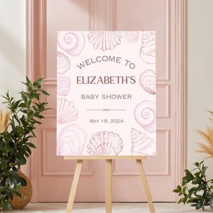 May include: A light pink welcome sign on a wooden easel, featuring seashell illustrations. The sign reads "Welcome to Elizabeth's Baby Shower" with the date "May 18, 2024". Decorative plants flank the sign.