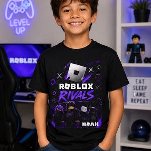 May include: A black short-sleeve t-shirt featuring a graphic print with the Roblox logo, the word Rivals, and four blocky character figures in purple and black tones. The design includes the name Noah in white text at the bottom of the graphic.