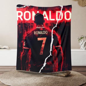 Puede incluir: Un tapiz rojo y negro con un jugador de f&uacute;tbol que lleva una camiseta negra con el nombre "RONALDO" y el n&uacute;mero "7" en la espalda. El jugador est&aacute; de pie frente a un fondo rojo y negro con la palabra "RONALDO" escrita en letras blancas.