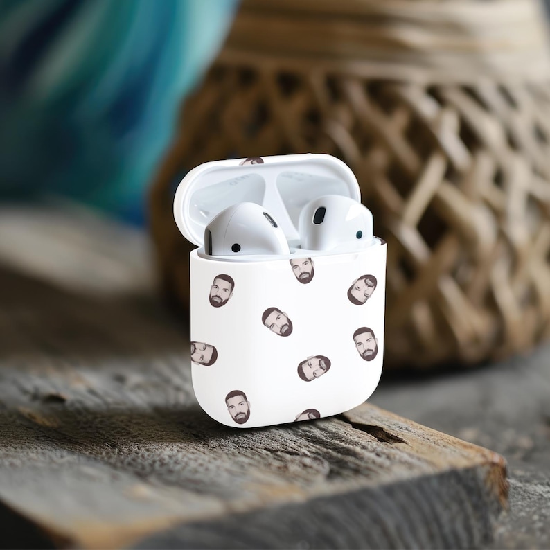 May include: White wireless headphone case with a repeating pattern of a person's face. The case is open and the headphones are visible inside.