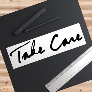 May include: A black rectangular surface with the words "Take Care" in black script. A silver ruler with inch measurements is on the right. Two black pens are at the top.