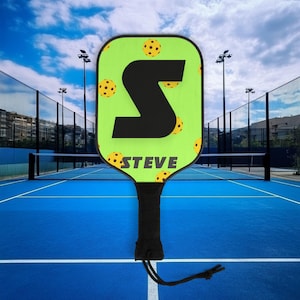 Könnte beinhalten: Ein grüner Pickleball-Schläger mit einem schwarzen Buchstaben "S" und dem Namen "STEVE" darauf gedruckt. Der Schläger hat einen schwarzen Griff und eine schwarze Schnur, die am unteren Ende befestigt ist.