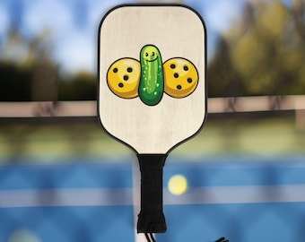 May include: A wooden pickleball paddle with a green pickle and two yellow pickleballs with black dots on it. The paddle has a black handle with a black string.