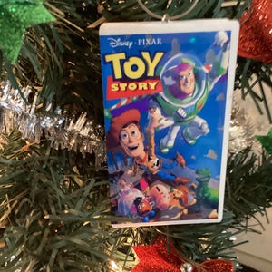 Może przedstawiać: Prostokątna ozdoba choinkowa z okładką filmu "Toy Story". Ozdoba przedstawia Buzza Astrala i Chudego z logo Disney Pixar. Ozdoba wisi na zielonej choince z czerwonymi i zielonymi błyszczącymi dekoracjami.