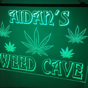 May include: Green LED sign with the text "Aidan's Weed Cave" in white lettering. The sign features several stylized marijuana leaf designs.