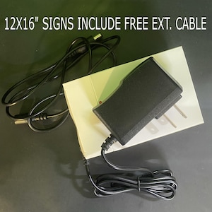 May include: A black 12V power supply with a black cord and a white plug. The power supply is on a white surface with the text "12X16" SIGNS INCLUDE FREE EXT. CABLE" printed on it.