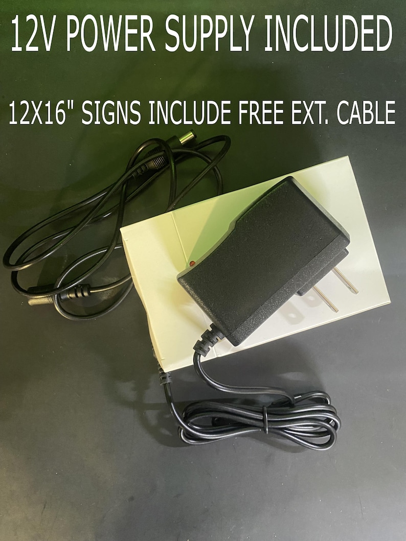 May include: A black 12V power supply with a black cord and a white plug. The power supply is on a white surface with the text "12X16" SIGNS INCLUDE FREE EXT. CABLE" printed on it.