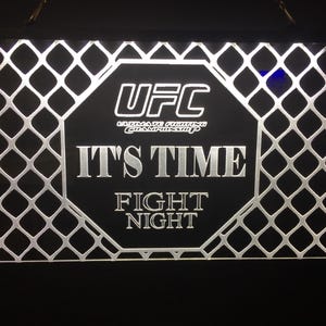May include: A black and white sign with a white octagon in the center. The octagon has the text "UFC Ultimate Fighting Championship" and "IT'S TIME FIGHT NIGHT". The sign is surrounded by a black and white cage pattern.