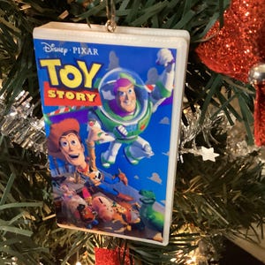 May include: A Christmas tree ornament shaped like a VHS tape of the Disney Pixar movie "Toy Story." The ornament features Buzz Lightyear and Woody on the cover, with the Disney Pixar logo. The ornament is hanging on a Christmas tree.