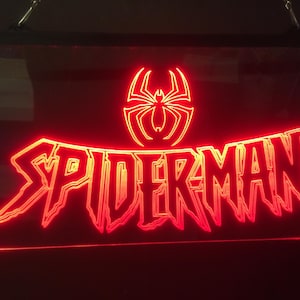 Può includere: Un cartello al neon rosso con la parola "SPIDERMAN" in un font stilizzato. Un logo di ragno rosso è sopra la parola.