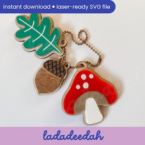 May include: A wooden keychain featuring an oak leaf, acorn, and mushroom charm. The leaf is green with white veins, the acorn is brown, and the mushroom has a red cap with white spots. The keychain includes the text "instant download • laser-ready SVG file".